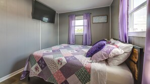 Un dormitorio con cama, cortinas moradas, una colcha y un televisor montado en la pared.