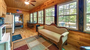 A cozy cabin interior with wooden walls, a sofa, a dining table, and large windows offering a view of the outdoors. - 3 Mi to Dale Hollow Lake: Wooded Retreat w/ Decks! (Byrdstown)