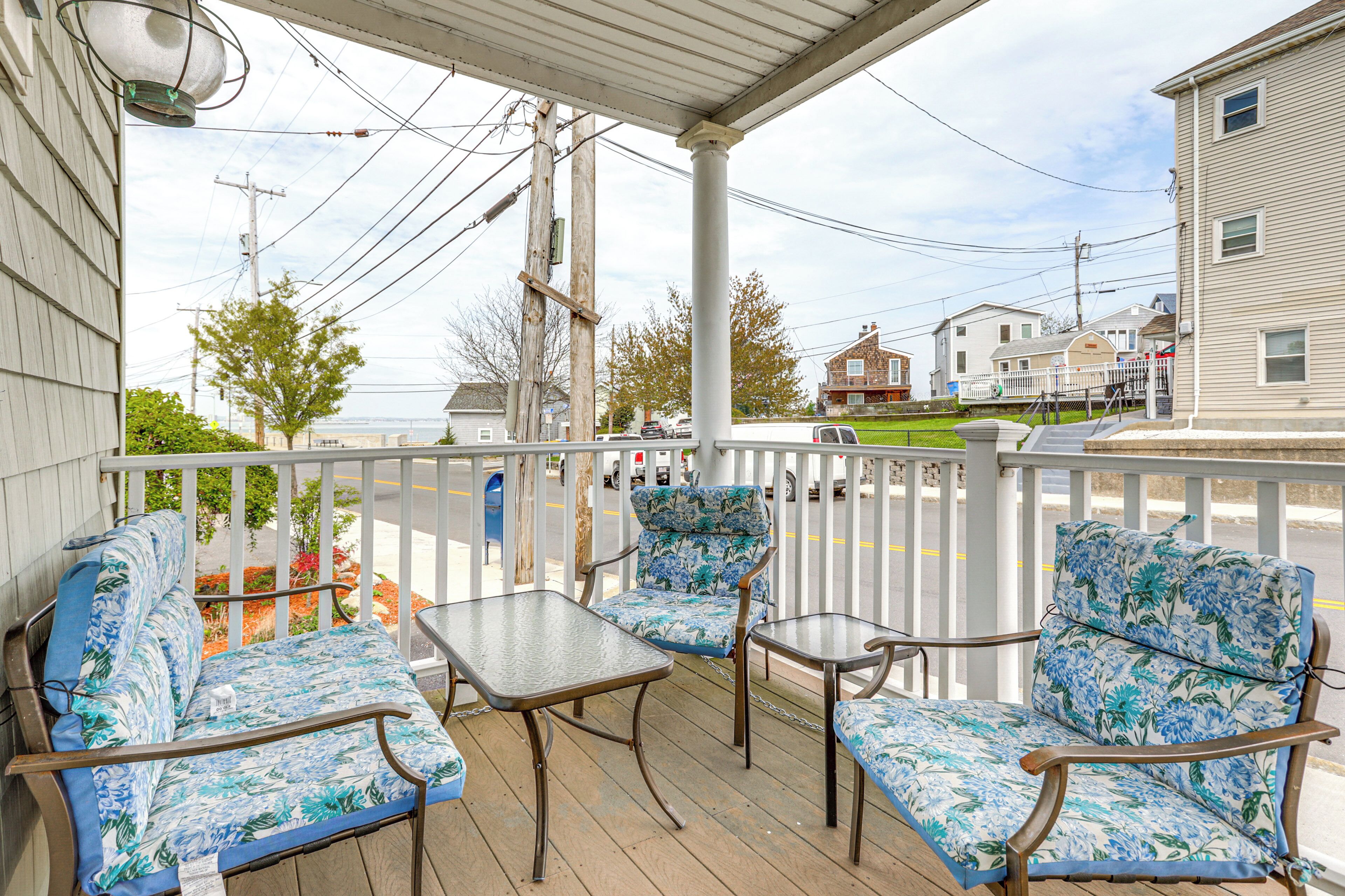 A covered porch with two chairs and a table, overlooking a street with houses and a view of the water.