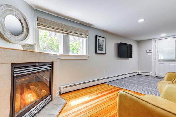 A modern living room with a fireplace, a large window with blinds, a flat-screen TV mounted on the wall, and a door leading to another room.