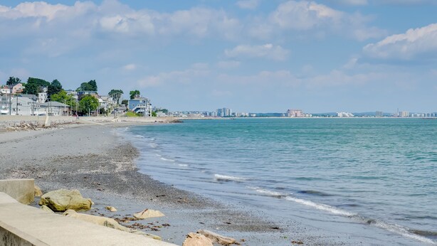 Une plage avec une promenade en béton, une eau calme et des bâtiments en arrière-plan.