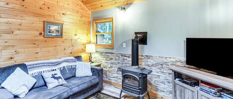 A cozy cabin interior with a wooden ceiling, a stone wall, a fireplace, a sofa, a television, and a cabinet with books.