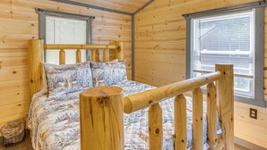 A wooden cabin bedroom with a bed, two windows, and a wooden railing.