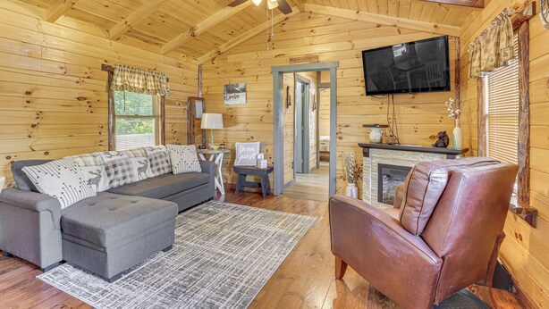 A cozy cabin interior with a sofa, ottoman, leather chair, television, fireplace, and a ceiling fan.