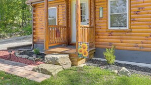 A log cabin with a wooden porch, a front door, and a small garden area with rocks and a flag.