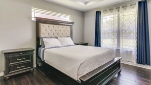 A bedroom with a tufted headboard, a wooden nightstand, a bed with a white bedspread, and a window with blue curtains. - Fenced Yard: Home < 1 Mi to Mayor Pablo Peña Park (Weslaco)