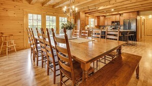 Een ruime houten eetkamer met een grote tafel en stoelen, een kroonluchter, en een keuken met houten kasten en apparatuur.