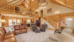 Een ruime woonkamer met een hoge plafond, houten balken, een trap, leren banken en een open haard.