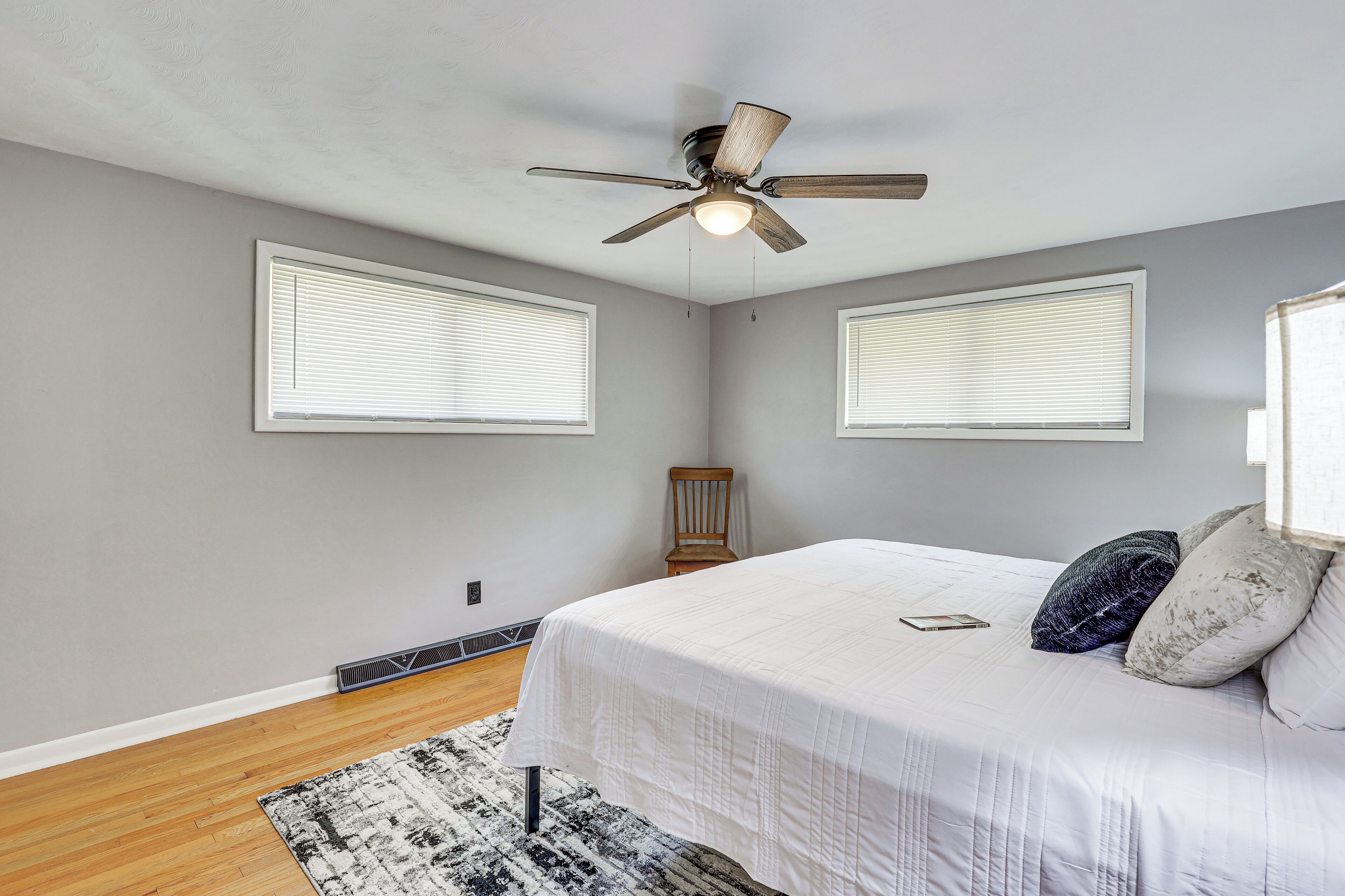 A bedroom with a bed, a ceiling fan, two windows with blinds, a chair, and a rug.