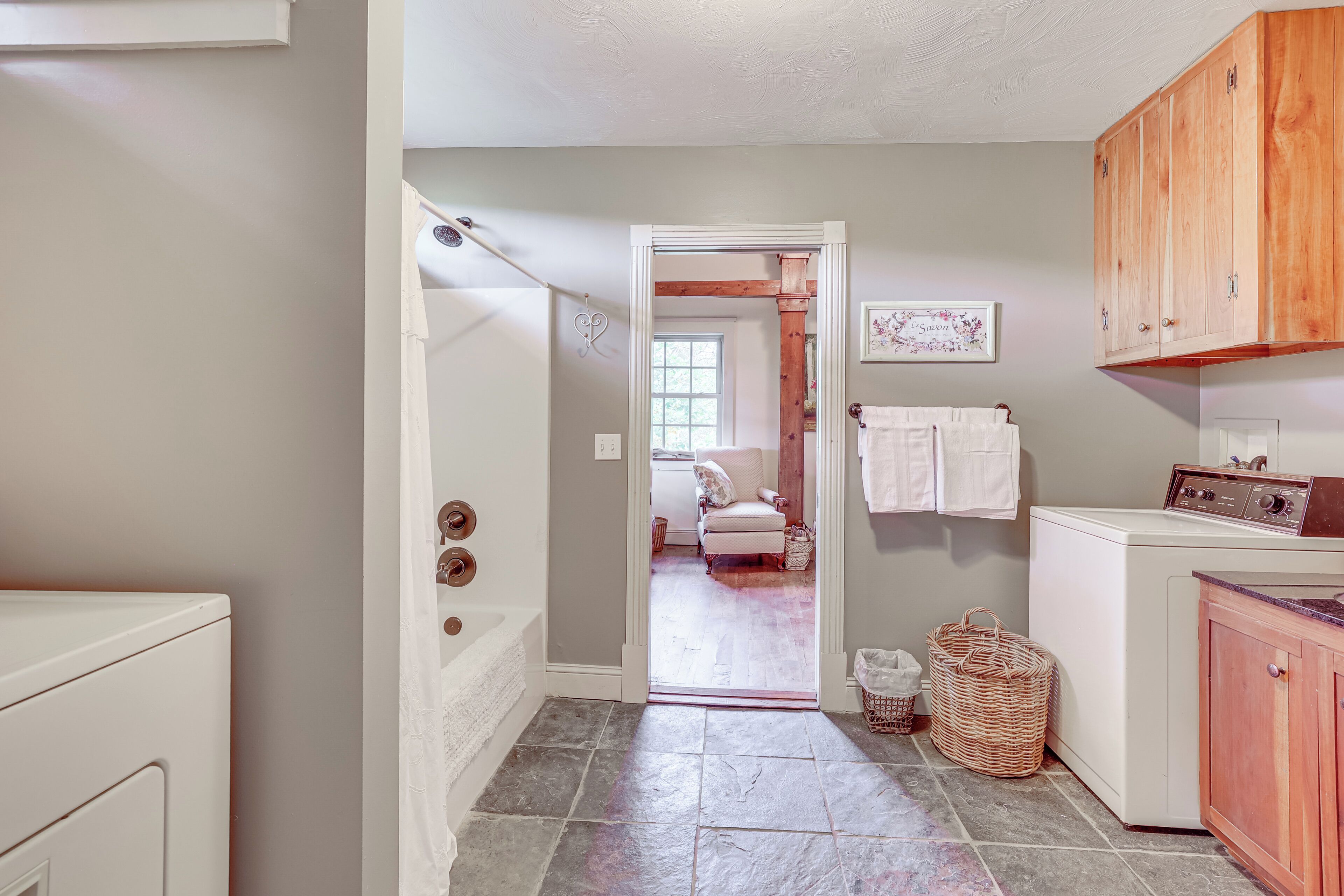 A laundry room with a washing machine, a dryer, and wooden cabinets. There is a door leading to a bedroom with a bed and a window.