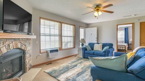 A living room with a stone fireplace, a blue sofa, a large window, and a ceiling fan. - Port Hope Escape: Fire Pit, Deck & Lake Views (Port Hope)