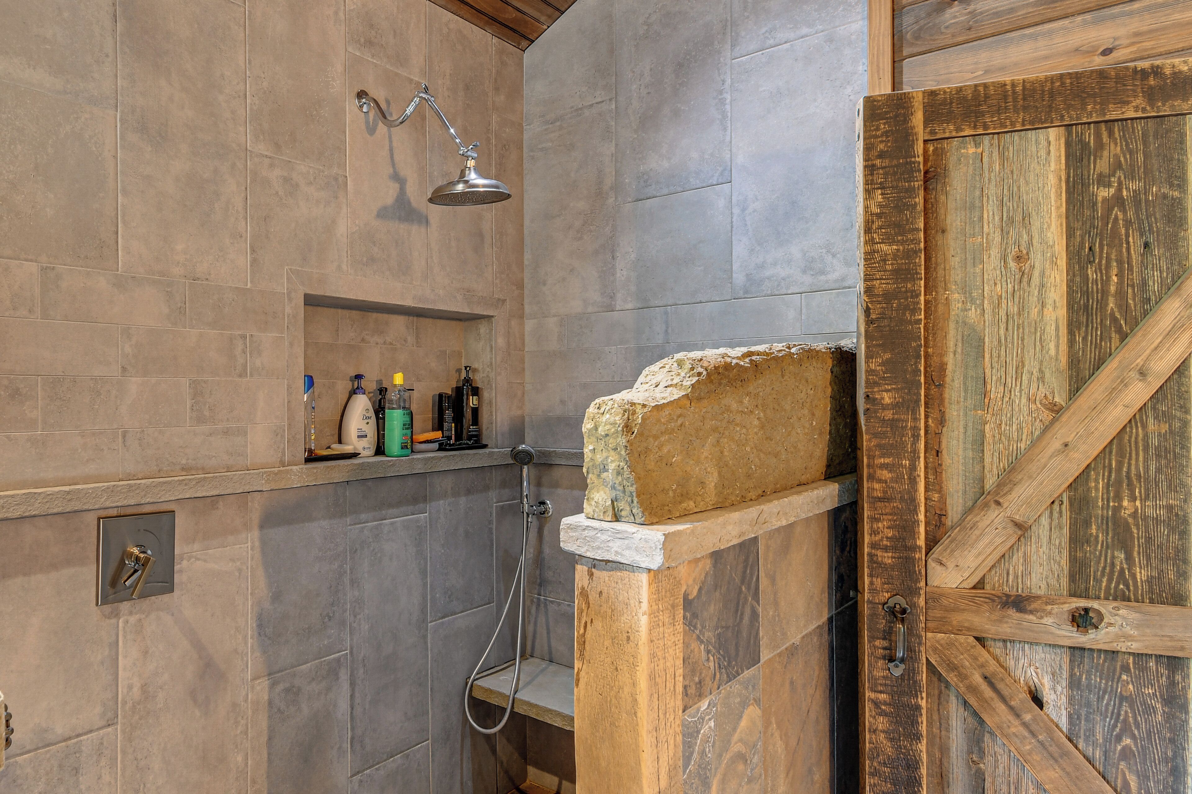 A bathroom with a shower, a wooden door, and a shelf with toiletries.