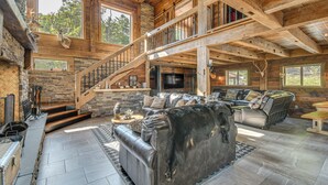 A spacious living room with a large leather sofa, a stone fireplace, and a wooden staircase. - Luxe Cabin Compound 4 Mi to Cameron State Forest! (Cameron)