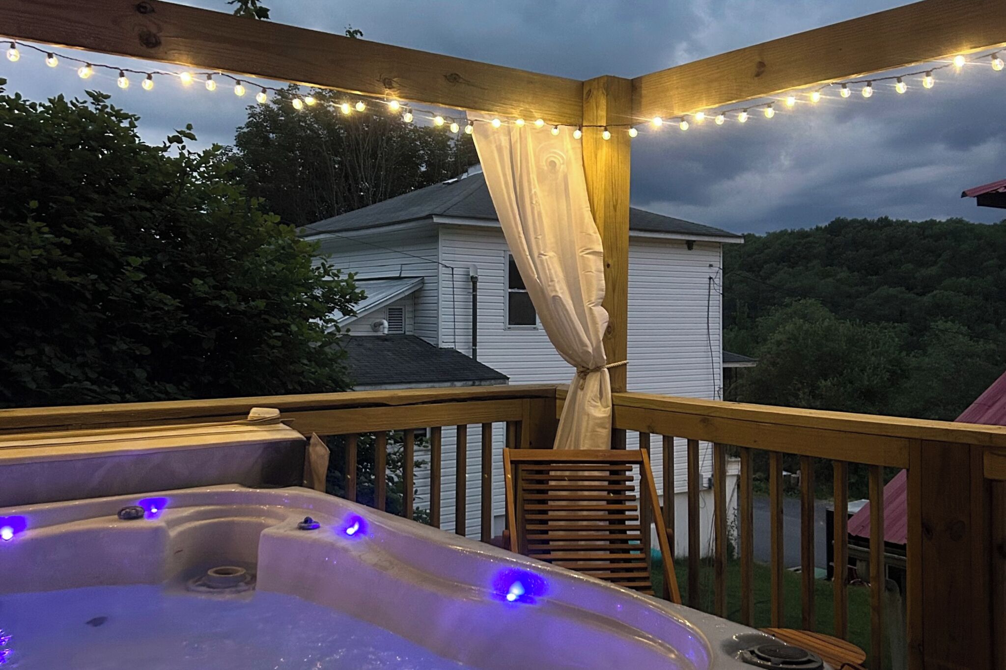 A wooden deck with a hot tub and string lights, overlooking a house and trees.