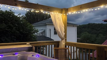 A wooden deck with a hot tub and string lights, overlooking a house and trees.
