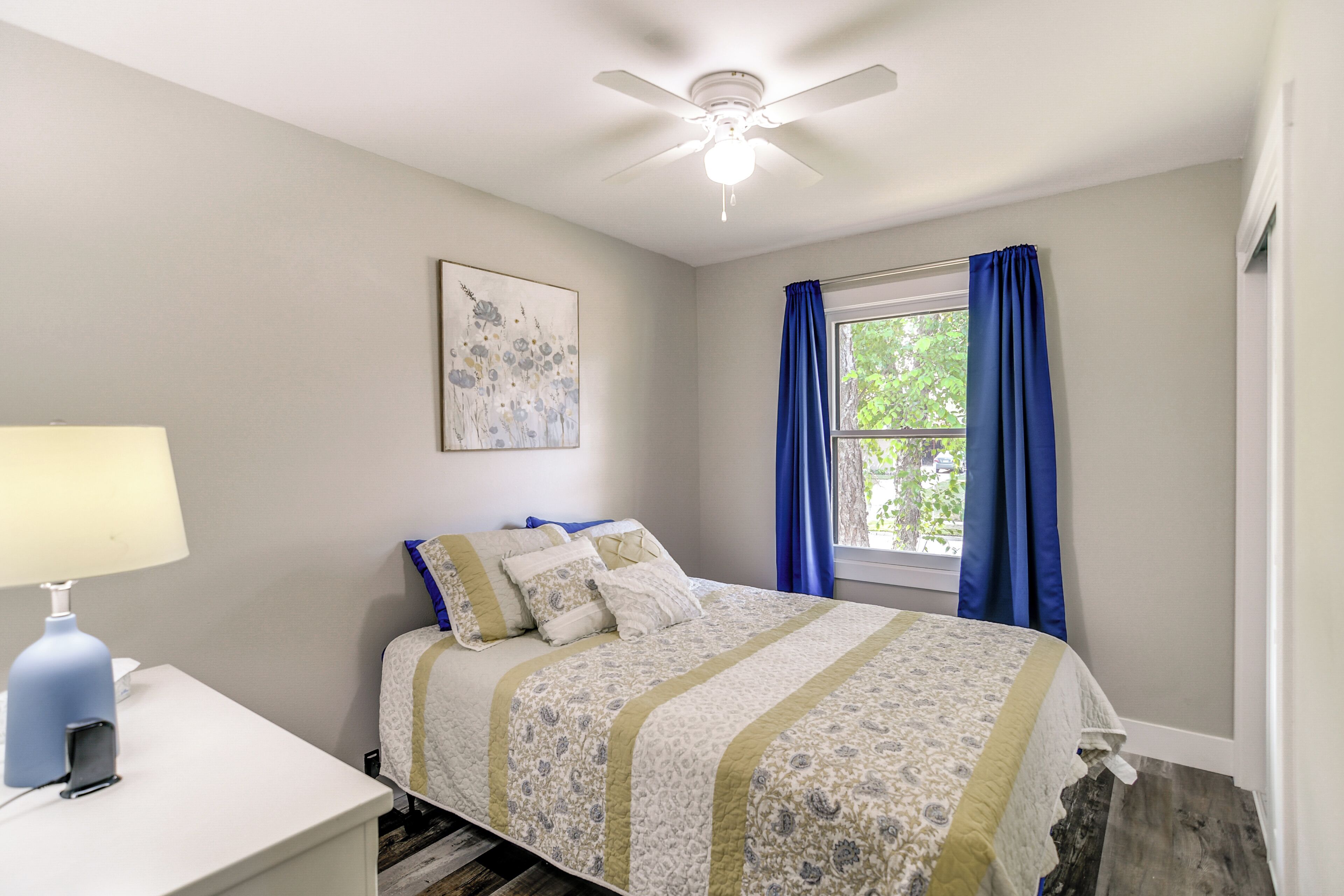 A bedroom with a bed, a ceiling fan, a window with blue curtains, and a wall-mounted lamp.