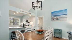A dining area with a wooden table and chairs, a kitchen in the background, and a painting of a beach scene on the wall. - 2D West Hyde Park - Your New Favorite Beach Villa (Myrtle Beach)