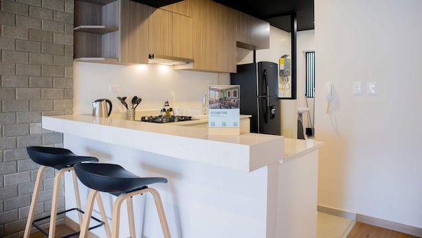 Une cuisine moderne avec un plan de travail blanc, deux tabourets de bar noirs, un réfrigérateur noir et des meubles de rangement en bois.