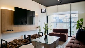 Un salon moderne avec une table basse en verre, un canapé, un téléviseur fixé au mur et une vue sur la ville grâce à de grandes fenêtres.