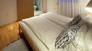 A bedroom with a wooden bed frame, a large window with curtains, a bedside lamp, and a wooden ceiling beam.