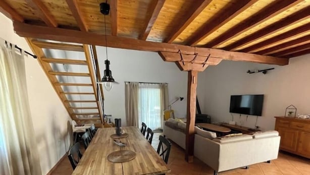 A spacious living area with a wooden dining table, sofa, and TV. A staircase leads to an upper level.
