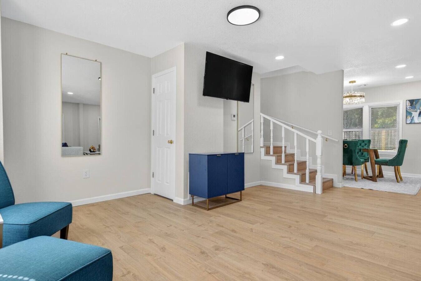 A modern living room with a blue sofa, a flat-screen TV mounted on the wall, a staircase leading to a dining area with green chairs, and a white door.