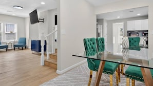 Ein modernes Wohnzimmer mit Treppe, einem Essbereich mit Glastisch und sesseln in Türkis und einer Küche mit weißen Schränken.