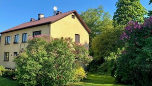 Een tweelaags huis met een schuin dak, schotelantenne en een verzorgde tuin met verschillende struiken en bloeiende planten.