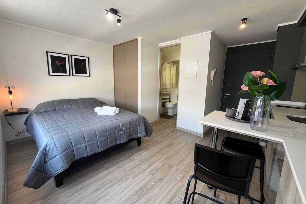 Une petite pièce comprenant un lit, un bureau et une chaise. On peut apercevoir l’espace douche à travers une porte ouverte.