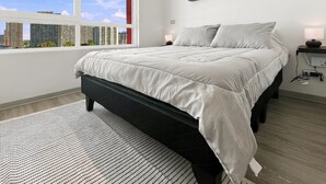 A modern bedroom with a large bed, a view of the city through a window, and a bedside table with a lamp. - Oceana Suites Vicuña Mackena (Santiago)