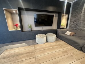 Egy modern nappali, melyben a falra van szerelve egy síkképernyős televízió, egy szürke kanapé, két kerek puff és egy fa padló található.