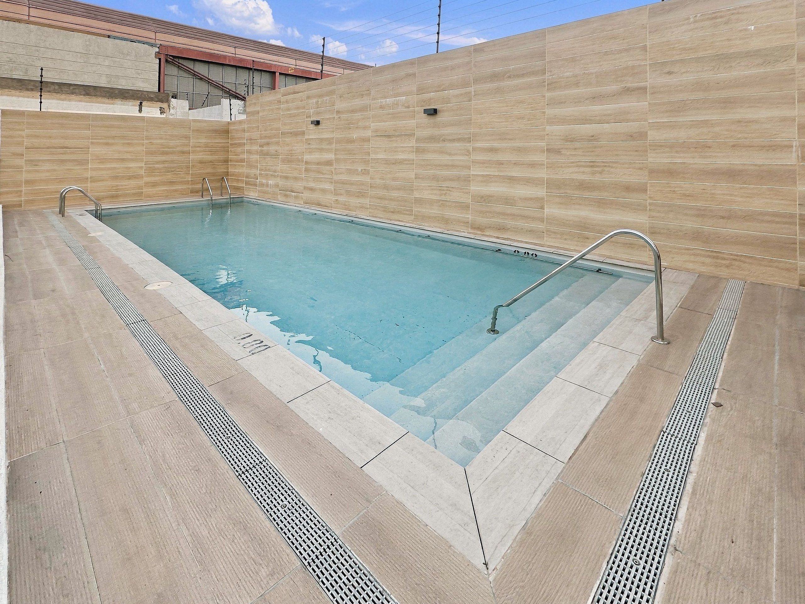 A rectangular swimming pool with a tiled deck, metal handrails, and a tiled wall surrounding it.