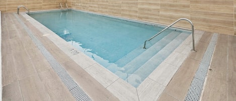 A rectangular swimming pool with a tiled deck, metal handrails, and a tiled wall surrounding it.