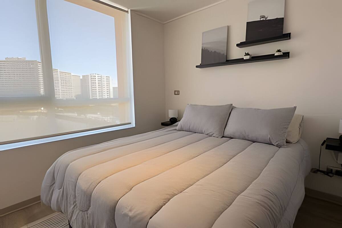 Une chambre avec une grande fenêtre, un lit recouvert d’une couette beige, deux tables de chevet et des étagères murales ornées d’objets décoratifs.