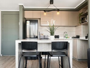 Een moderne keuken met een roestvrijstalen koelkast, twee barkrukken, een potplant en een koffiezetapparaat op het aanrecht.