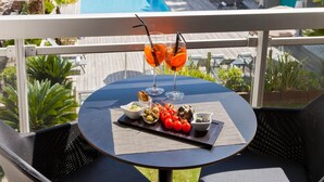 A balcony with a table set for two, overlooking a swimming pool and lounge area. - Pool View Junior Suite With Terrace (Cannes)
