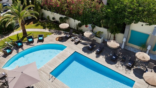 A large outdoor swimming pool with multiple lounge areas and umbrellas, surrounded by greenery and a white wall. - Pool View Junior Suite With Terrace (Cannes)