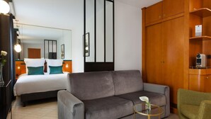 A modern hotel room with a grey sofa, a small round table with a glass top, a green armchair, a bed with white bedding, and a wooden wardrobe. - Pool View Junior Suite With Terrace (Cannes)