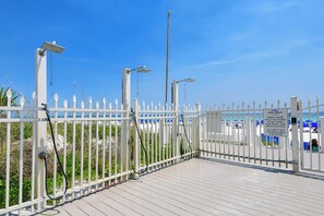 Un área de playa cercada con valla de piquetes blancos, un letrero que indica la necesidad de pulsera y llave de seguridad, y un cielo azul despejado.