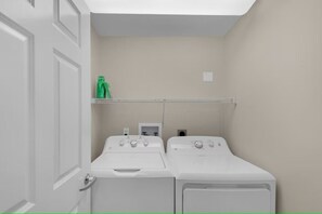 Uma lavanderia com duas máquinas de lavar e secar brancas de abertura frontal, uma prateleira com garrafas verdes e uma porta branca.