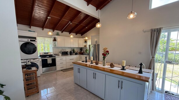 Una cocina amplia con armarios blancos, encimera de madera y una isla grande. Hay una zona de comedor con mesa y sillas, y vistas al exterior a través de un ventanal.