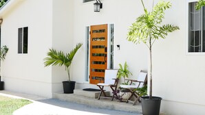 Un edificio blanco con puerta de madera, dos macetas y dos sillas blancas afuera.