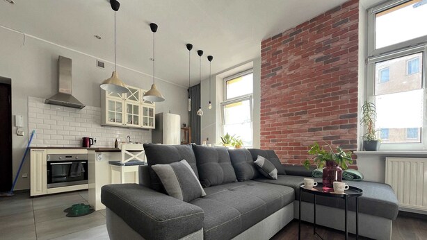 A modern living room with a grey sofa, a small side table, and a kitchen area with white cabinets and a brick wall.