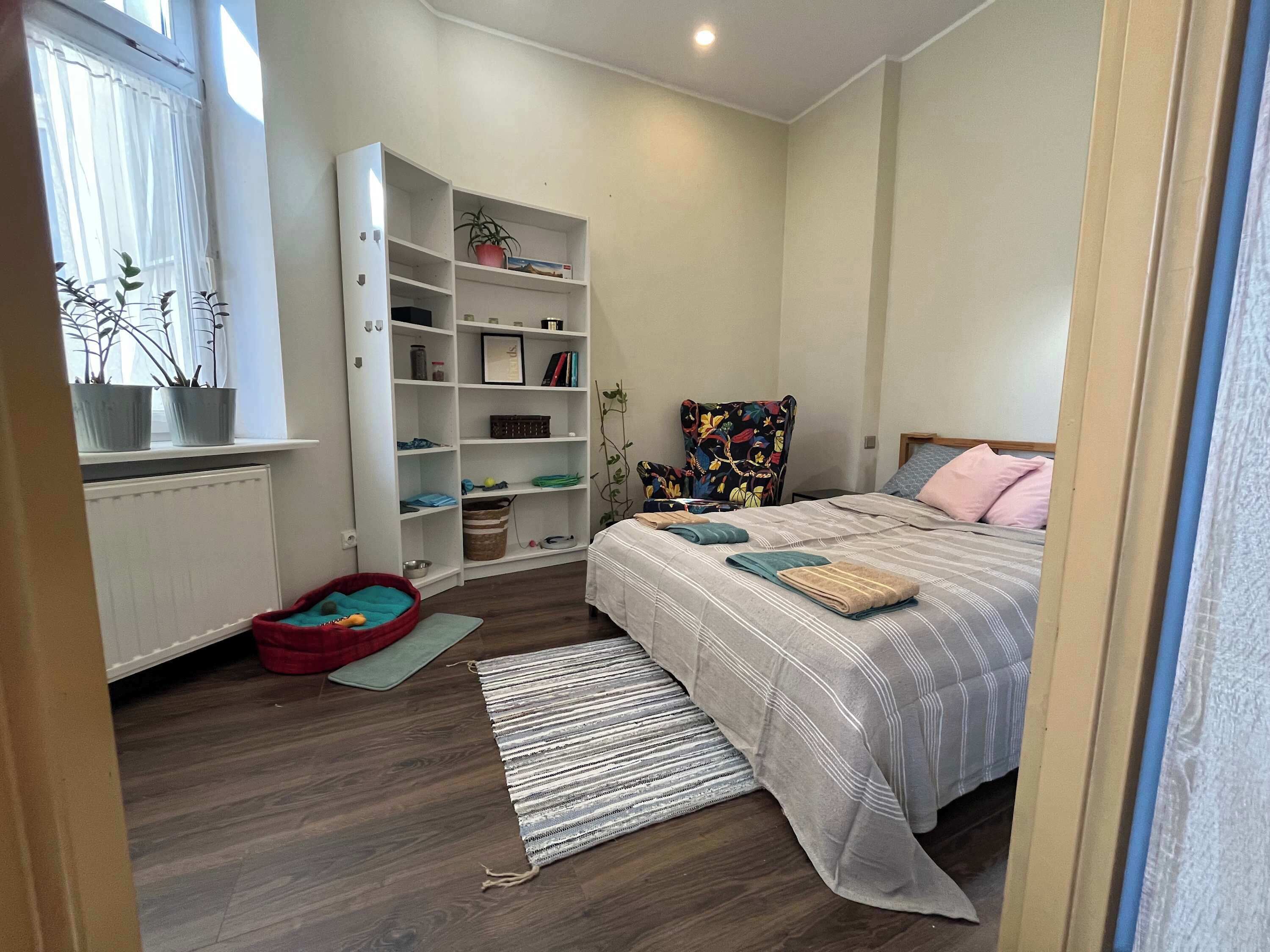 Un dormitorio con una cama, una estantería empotrada, una cama roja para mascotas, una alfombra pequeña y una ventana con cortinas.