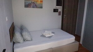 A small, single-bed room with a white mattress, two pillows, a folded towel, a painting of a sunset, a door, and a small shelf with a television.