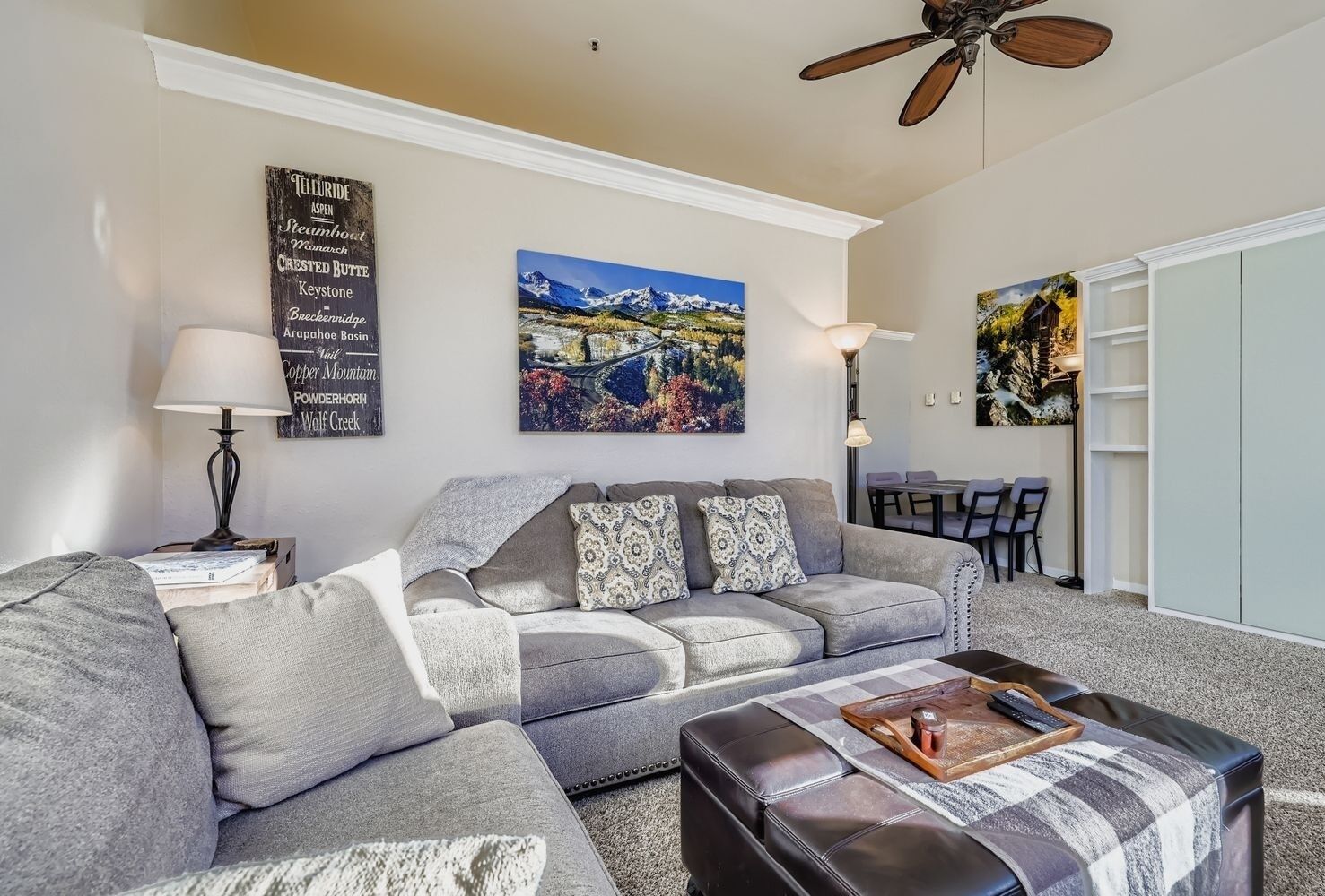 A modern living room with a grey sectional sofa, a black leather ottoman, and a painting of a mountain landscape.