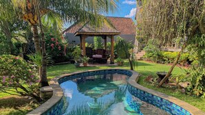 A swimming pool with blue mosaic tiles, surrounded by lush greenery and a gazebo.