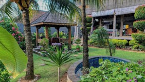 A garden with a gazebo, palm trees, a small pool, and a building with a tiled roof.