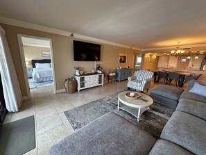 A living room with a sofa, armchair, coffee table, TV, and a kitchen area in the background.