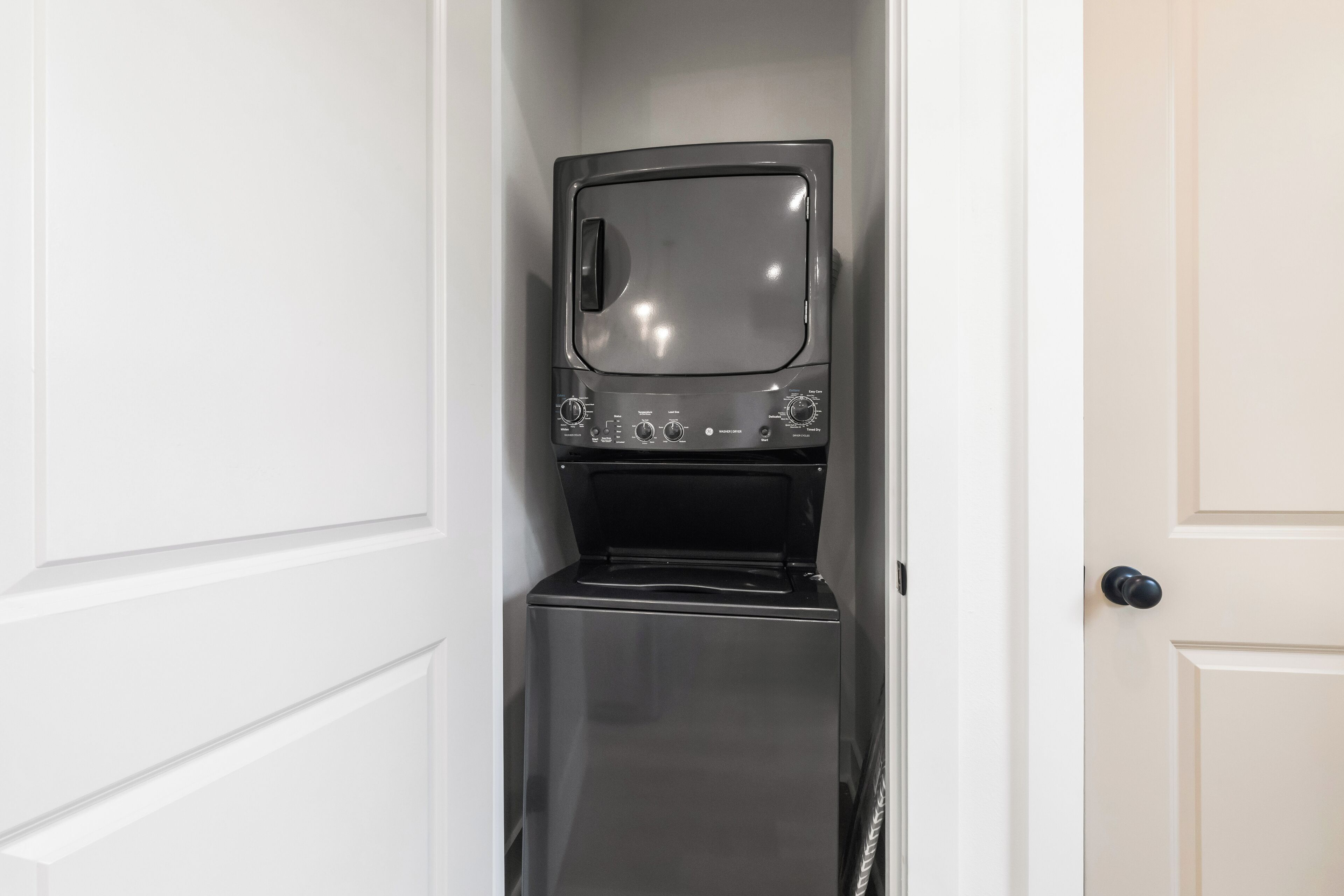 Un electrodoméstico compacto de acero inoxidable, probablemente una lavadora y secadora, en un estrecho pasillo con paredes blancas y una puerta a la derecha.